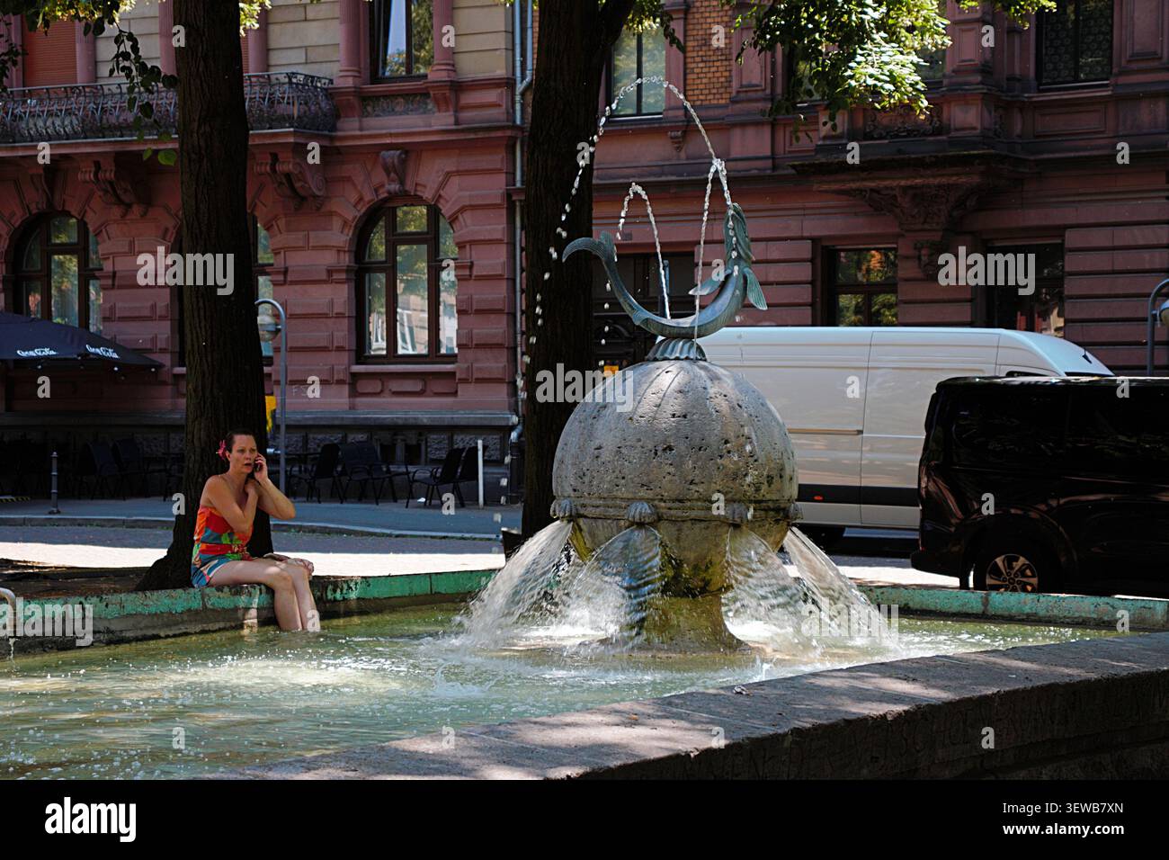 MAGONZA, RENANIA-PALATINATO, GERMANIA - 25 GIUGNO 2026: Donna che si raffredda i piedi nell'acqua presso la storica fontana di Fischtorbrunnen a Magonza, Germania. Foto Stock