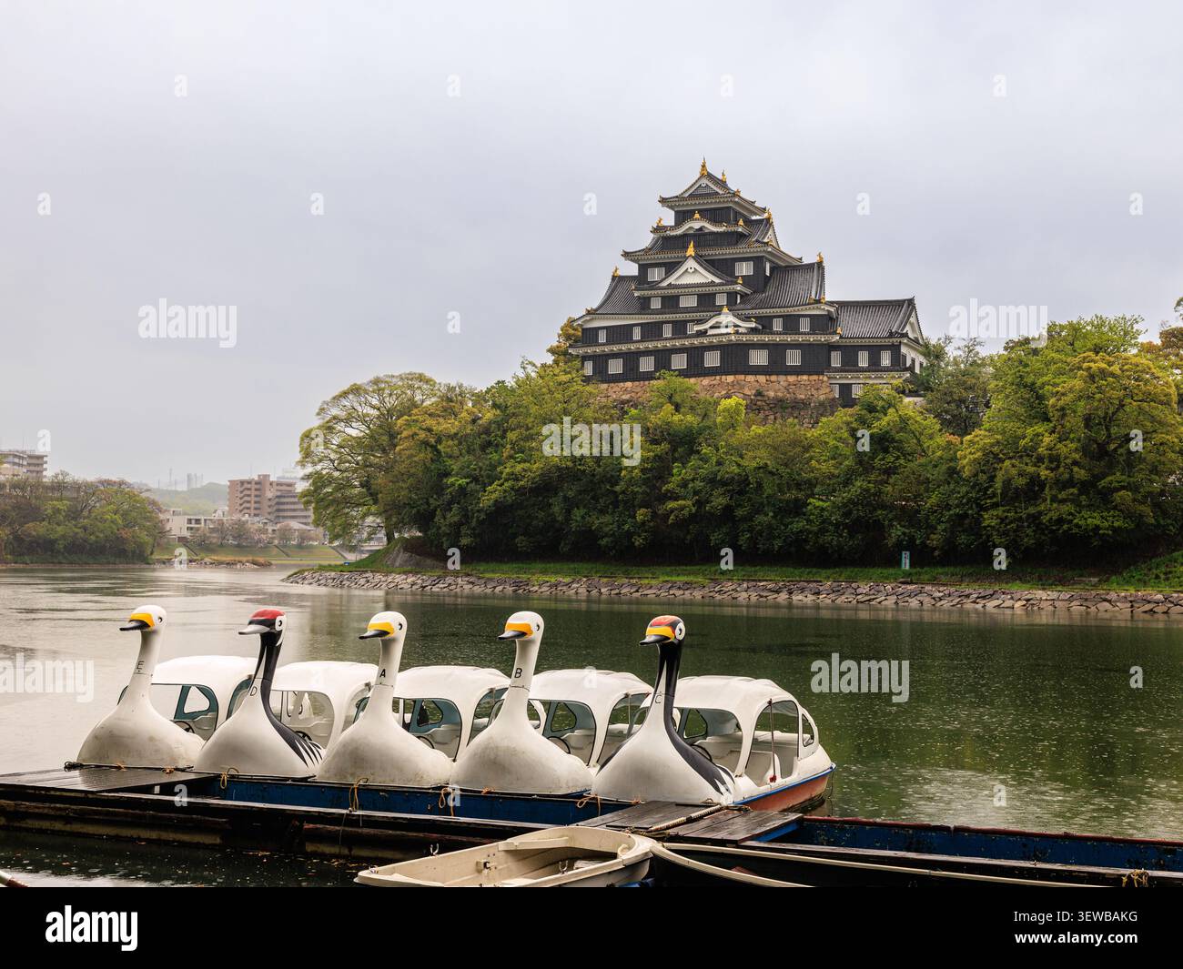 Pedalo a tema gru corona rossa attendono sul calmo fiume Asahi davanti all'imponente torrione nero del castello di Okayama Foto Stock