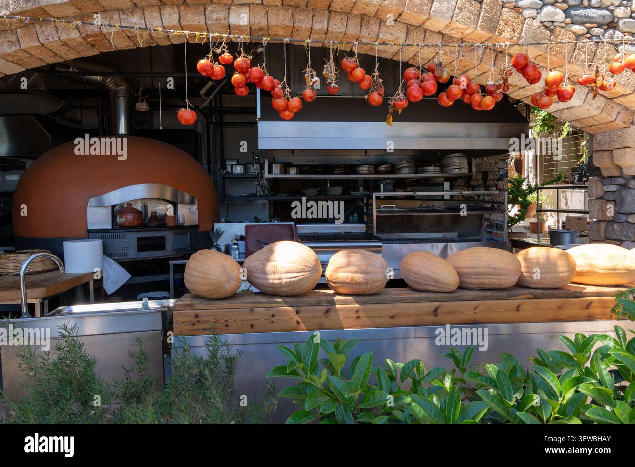Cucina rustica mediterranea sotto un arco di pietra con forno a legna per pizza, una fila di grandi zucche su un banco di legno e fasci Foto Stock