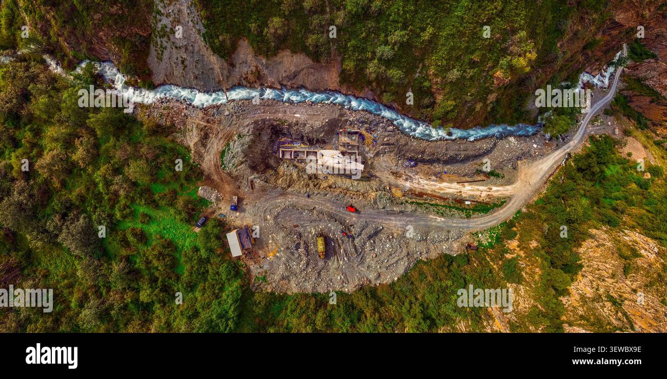 La vista aerea cattura un cantiere idroelettrico in un'aspra valle di montagna. Un vivace fiume turchese scorre lungo il sito, caratterizzato da una u Foto Stock