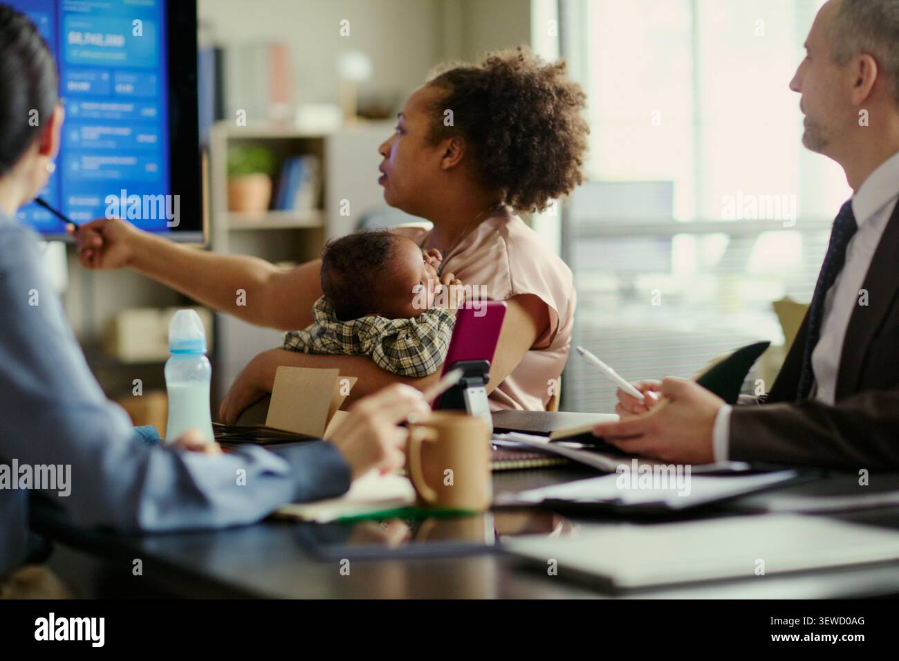 Giovane madre afroamericana che tiene in braccio il bambino durante la presentazione durante l'incontro d'ufficio con i colleghi, mostrando l'equilibrio dei genitori. Ideale per il congedo parentale, l'inclusione, il marketing di lavoro ibrido Foto Stock