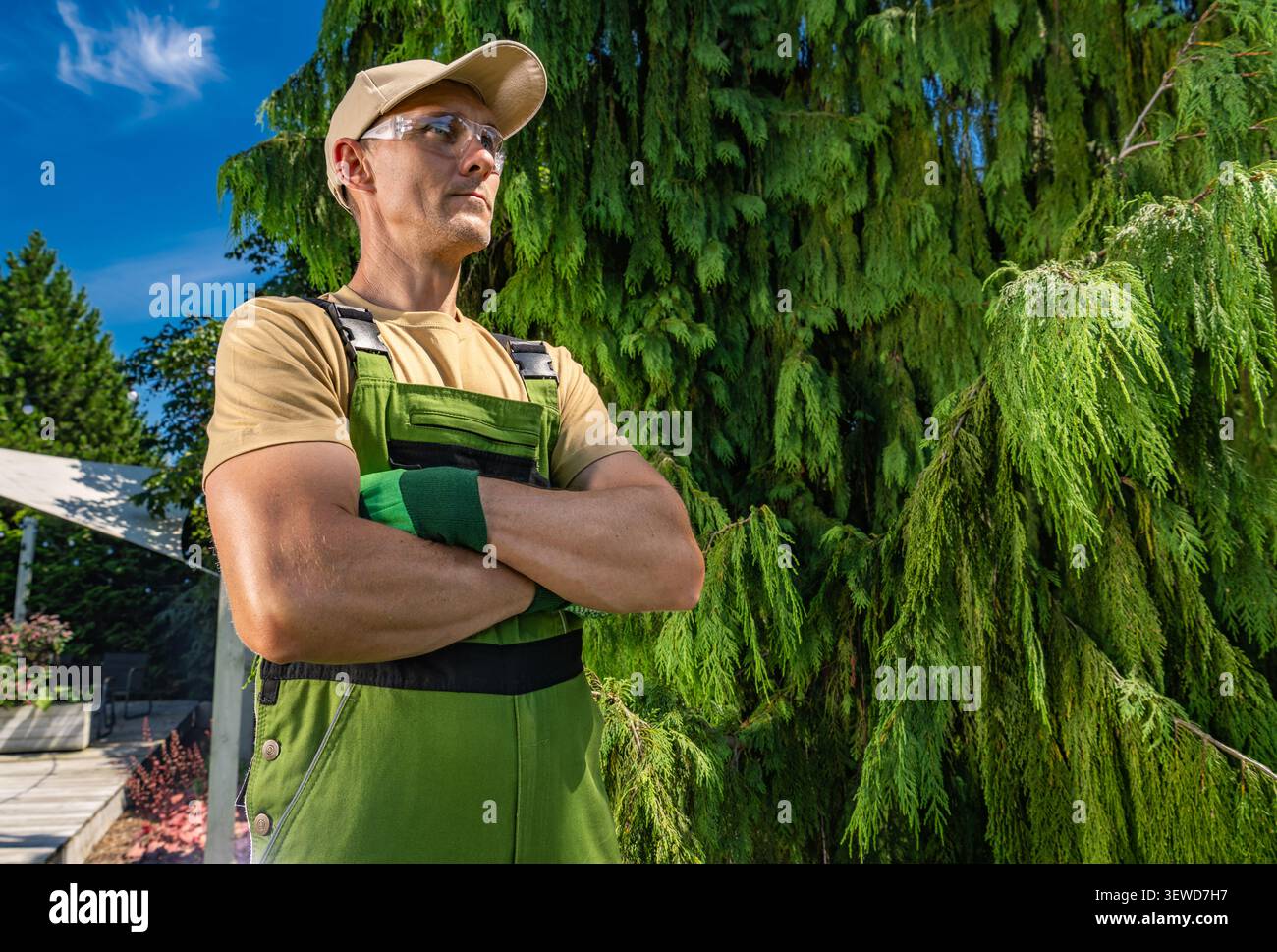 Un uomo con una camicia marrone e una tuta verde si staglia con le braccia incrociate. Guarda in lontananza mentre si trova vicino a un grande albero verde. Il sole Foto Stock