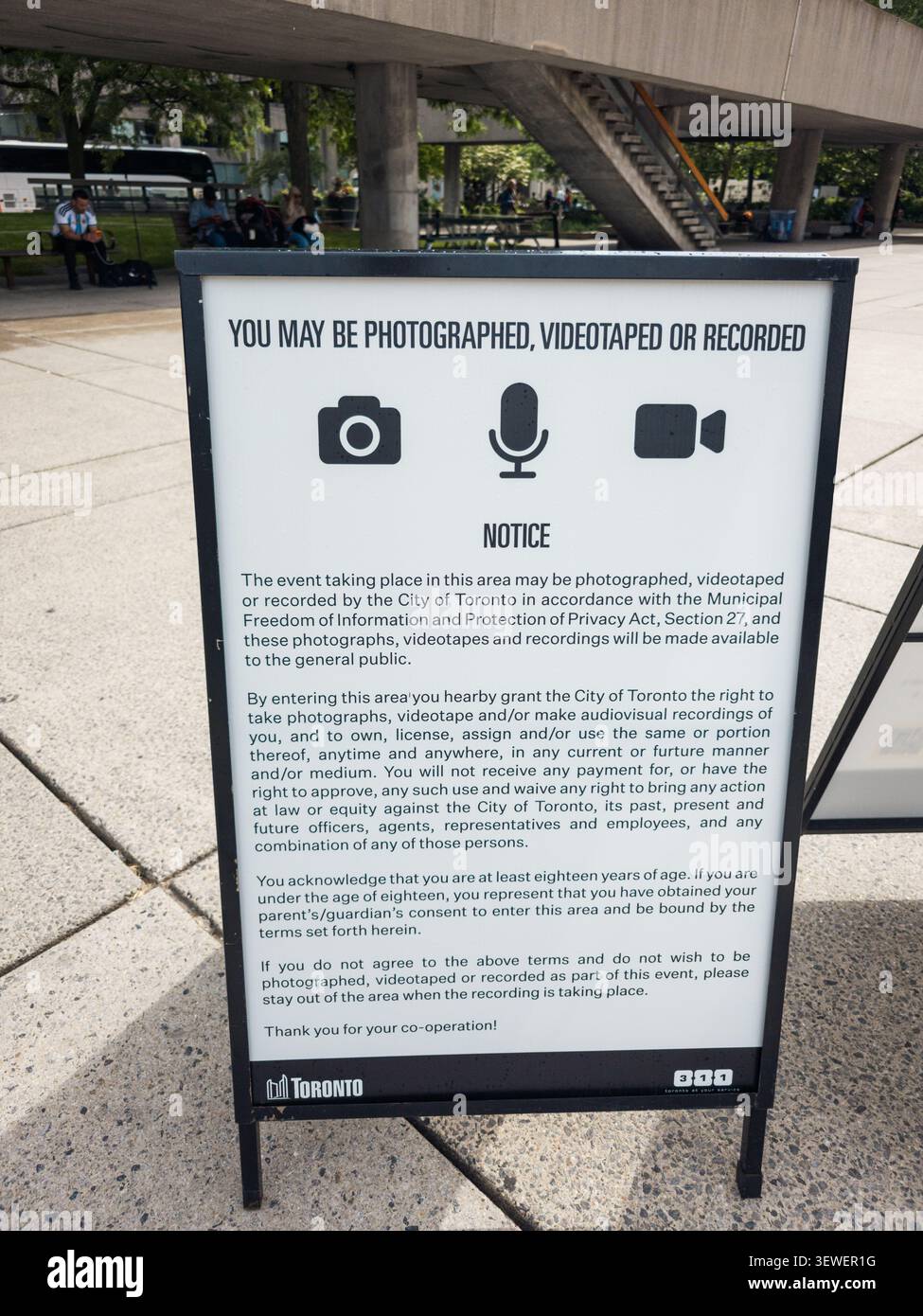 Un cartello di avviso della Città di Toronto informa i visitatori che nella zona possono avvenire fotografie, video e registrazioni audio. Persone sedute e camminano nelle vicinanze di un autobus - Immagine stock catturata con smartphone