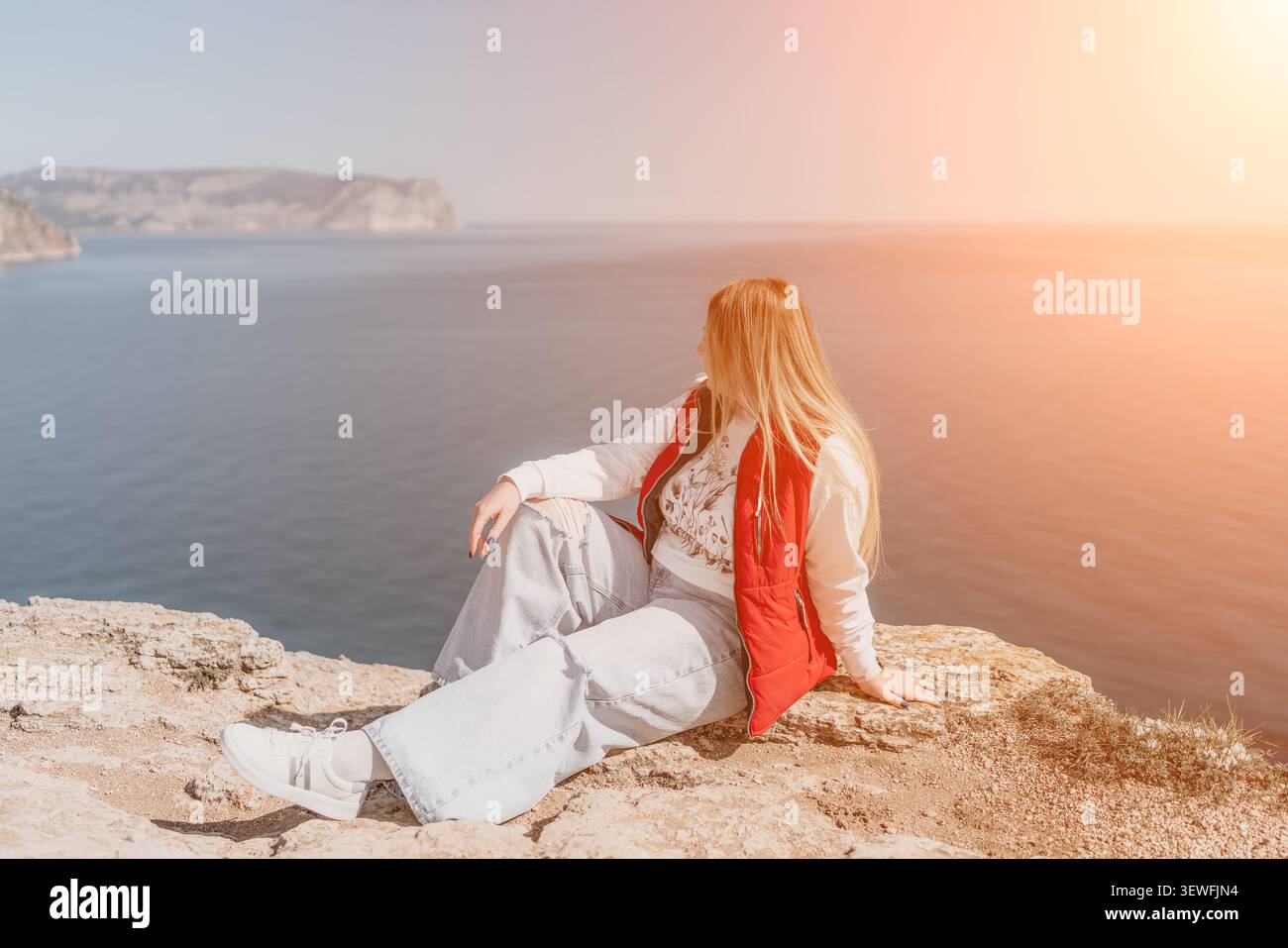 Woman Cliff Sea Bionda donna in un gilet rosso seduta sul bordo di una scogliera rocciosa che si affaccia sull'oceano durante l'ora d'oro Foto Stock