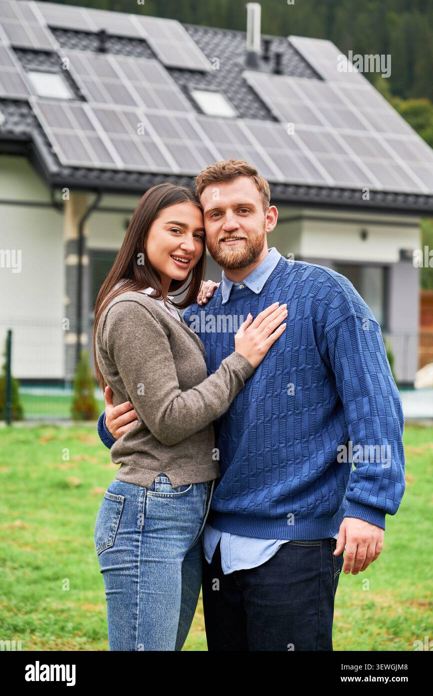 Coppia che abbraccia la casa vicino con pannelli solari, sorridendo alla macchina fotografica. Proprietari di case felici che utilizzano energia rinnovabile, casa ecocompatibile e concetto di stile di vita sostenibile Foto Stock
