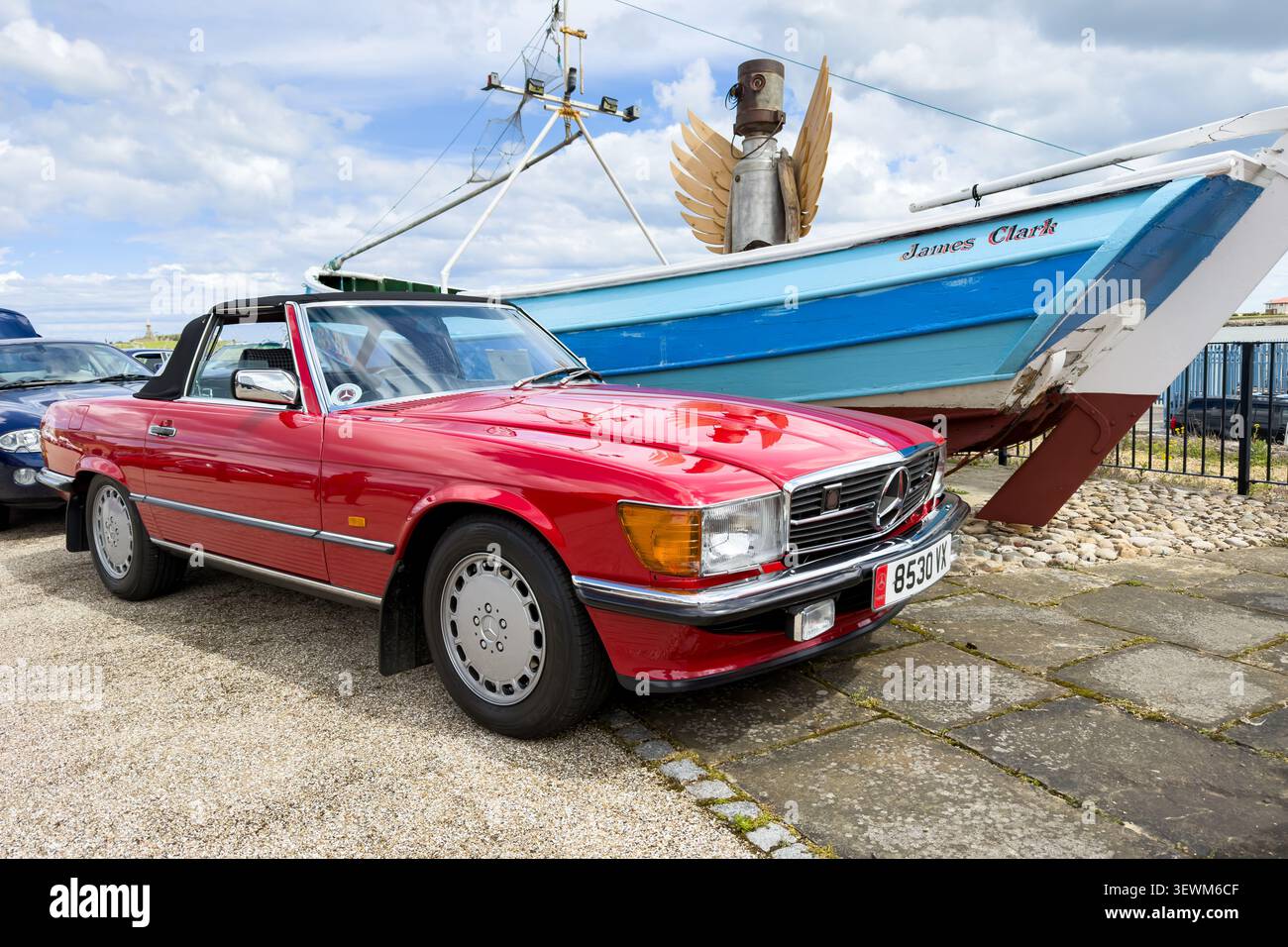 Mercedes-Benz SL R107 rossa e la barca James Clark parcheggiate all'aperto sotto un cielo nuvoloso Foto Stock