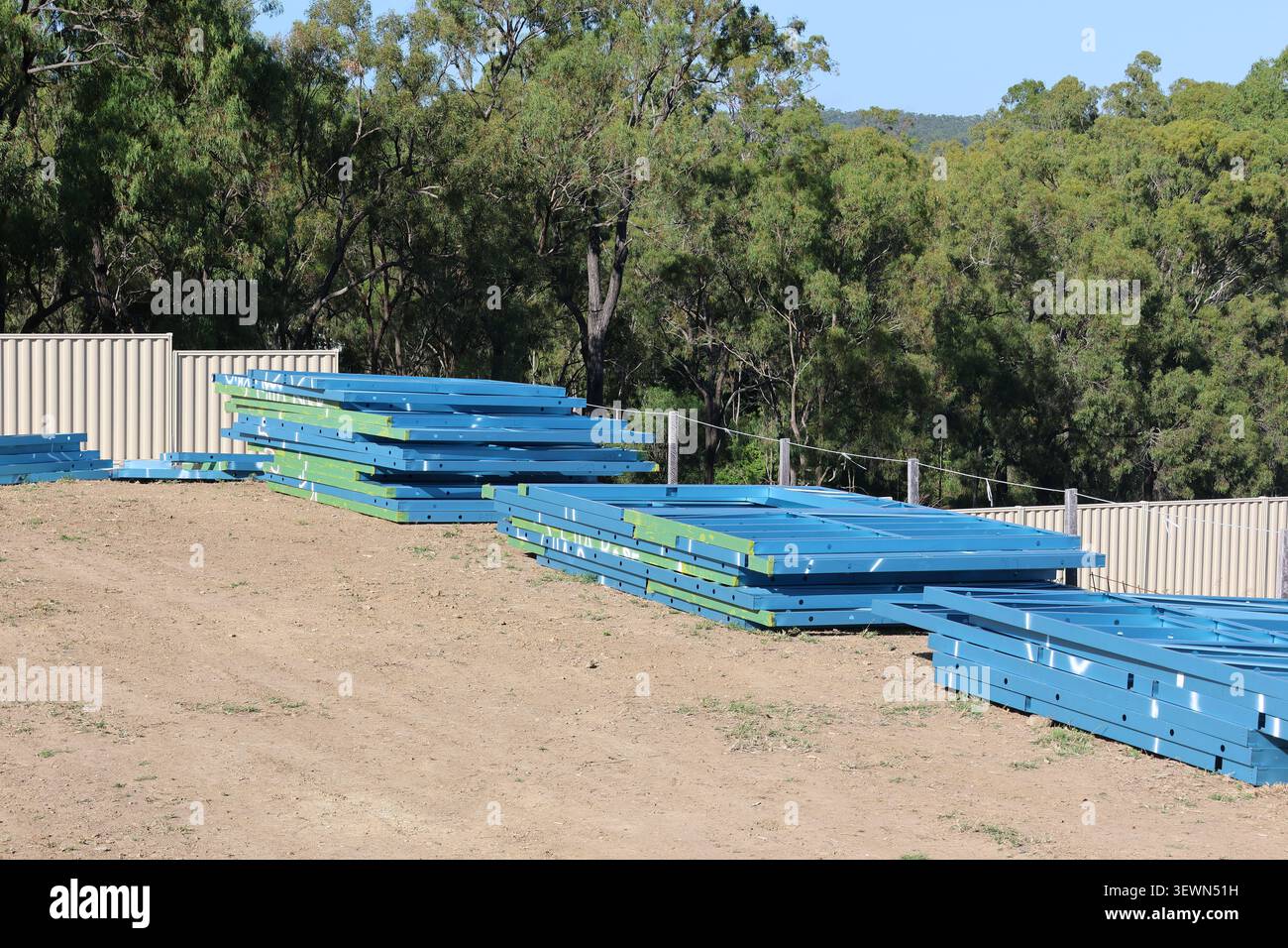 Pile di telai di case in acciaio blu conservate all'aperto su un terreno sterrato vicino agli alberi in un cantiere residenziale Foto Stock