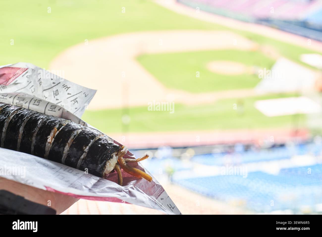 Involtini di riso alle alghe coreane a fette tenuti in mano contro un campo sportivo sfocato e posti a sedere blu dello stadio che mostrano un classico spuntino all'aperto Foto Stock