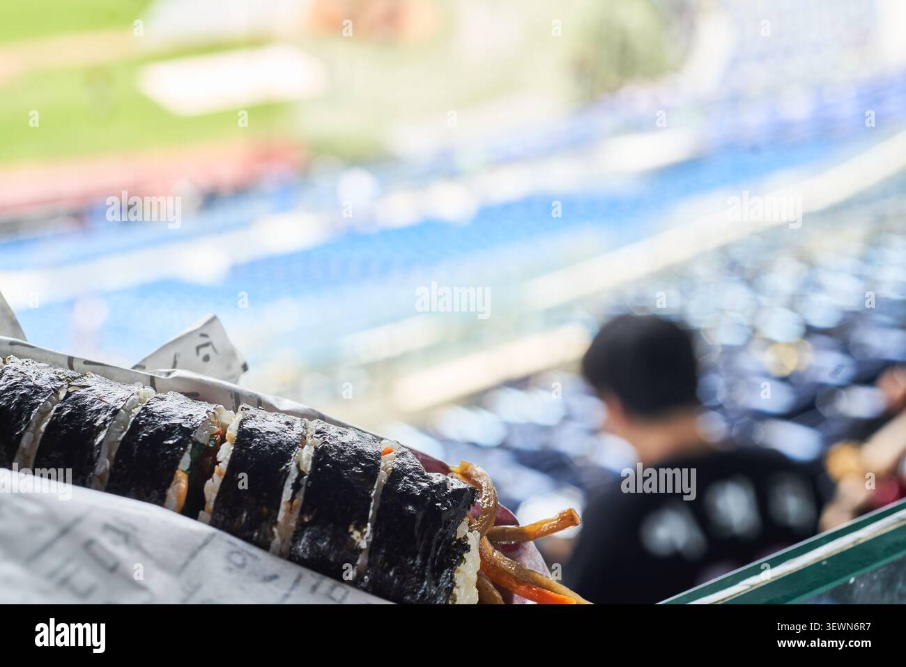 Involtini di riso alle alghe coreane a fette tenuti in mano contro un campo sportivo sfocato e posti a sedere blu dello stadio che mostrano un classico spuntino all'aperto Foto Stock