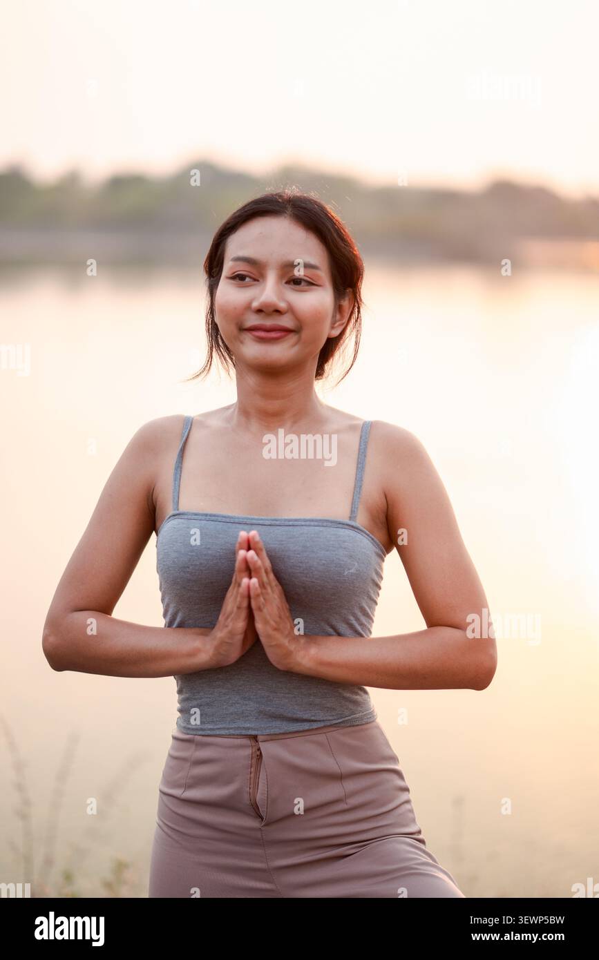 Ritratto di donna asiatica in posa di yoga sugli alberi con le mani di preghiera vicino a un lago calmo al tramonto, benessere tranquillo, equilibrio consapevole, fitness all'aperto, relax nella natura Foto Stock
