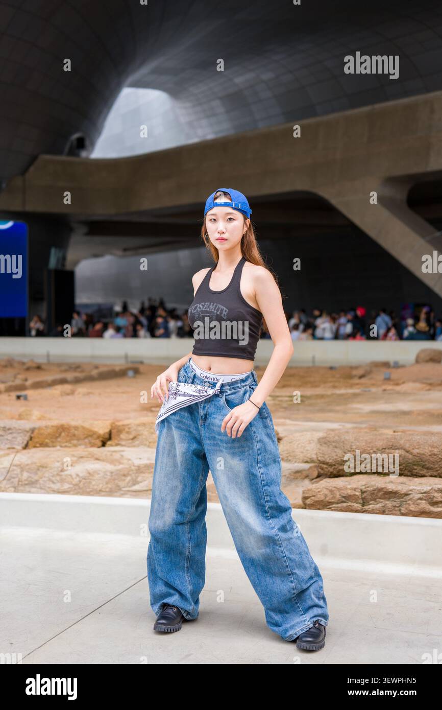Giovane donna asiatica elegante in jeans larghi blu, top corto nero e cappellino al contrario in posa all'aperto a Seoul, Corea del Sud. Foto Stock