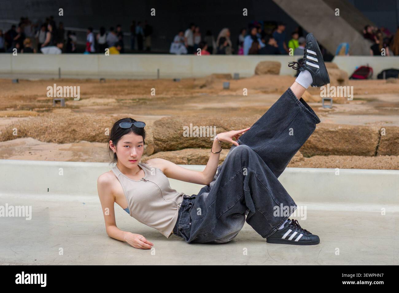 Giovane donna coreana che posa a terra con una gamba sollevata, indossando jeans oversize e scarpe da ginnastica durante uno shooting di moda all'aperto. Foto Stock