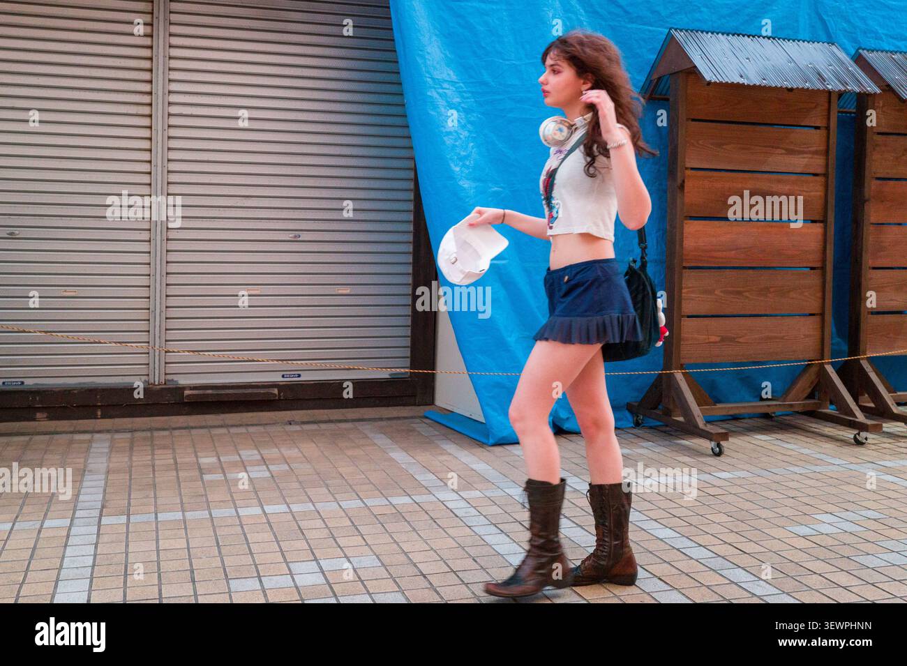 Giovane donna che cammina all'aperto con cuffie, crop top e stivali davanti a uno sfondo blu e una serranda chiusa. Foto Stock