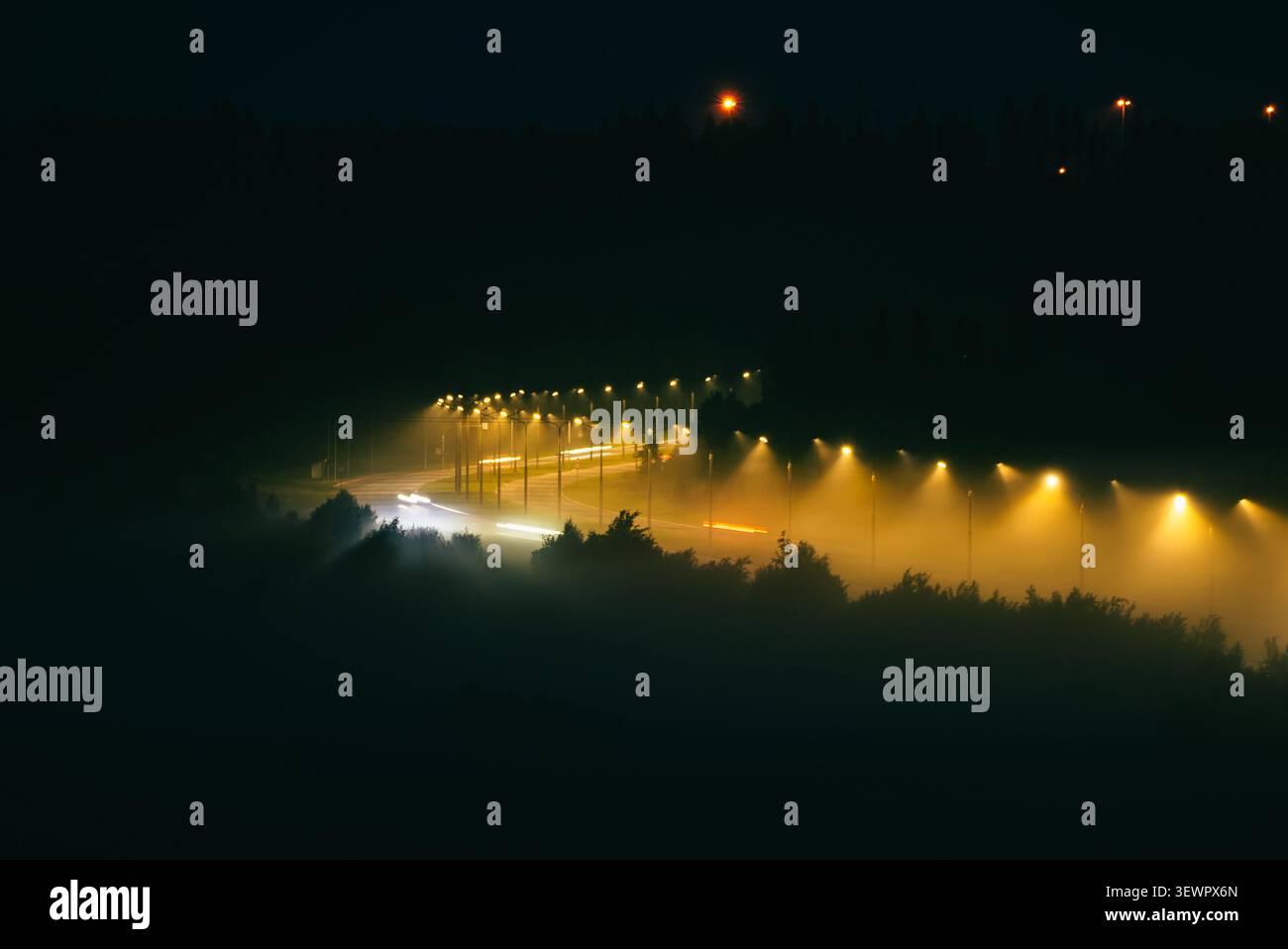 Autostrada avvolta dalla fitta nebbia di notte. File di potenti lampioni che illuminano l'atmosfera nebulosa. Mentre i fari e le luci di posizione posteriori sfocati si estendono lungo l'asfalto tortuoso Foto Stock