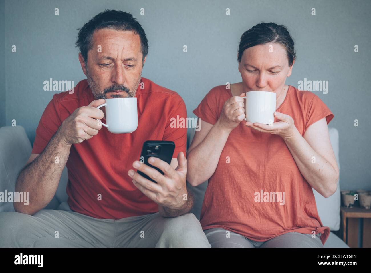 Una coppia di mezza età si siede insieme su un divano a casa, ognuno con una tazza di caffè mentre l'uomo fa scorrere il suo smartphone. Scena mattutina informale e rilassata su Foto Stock