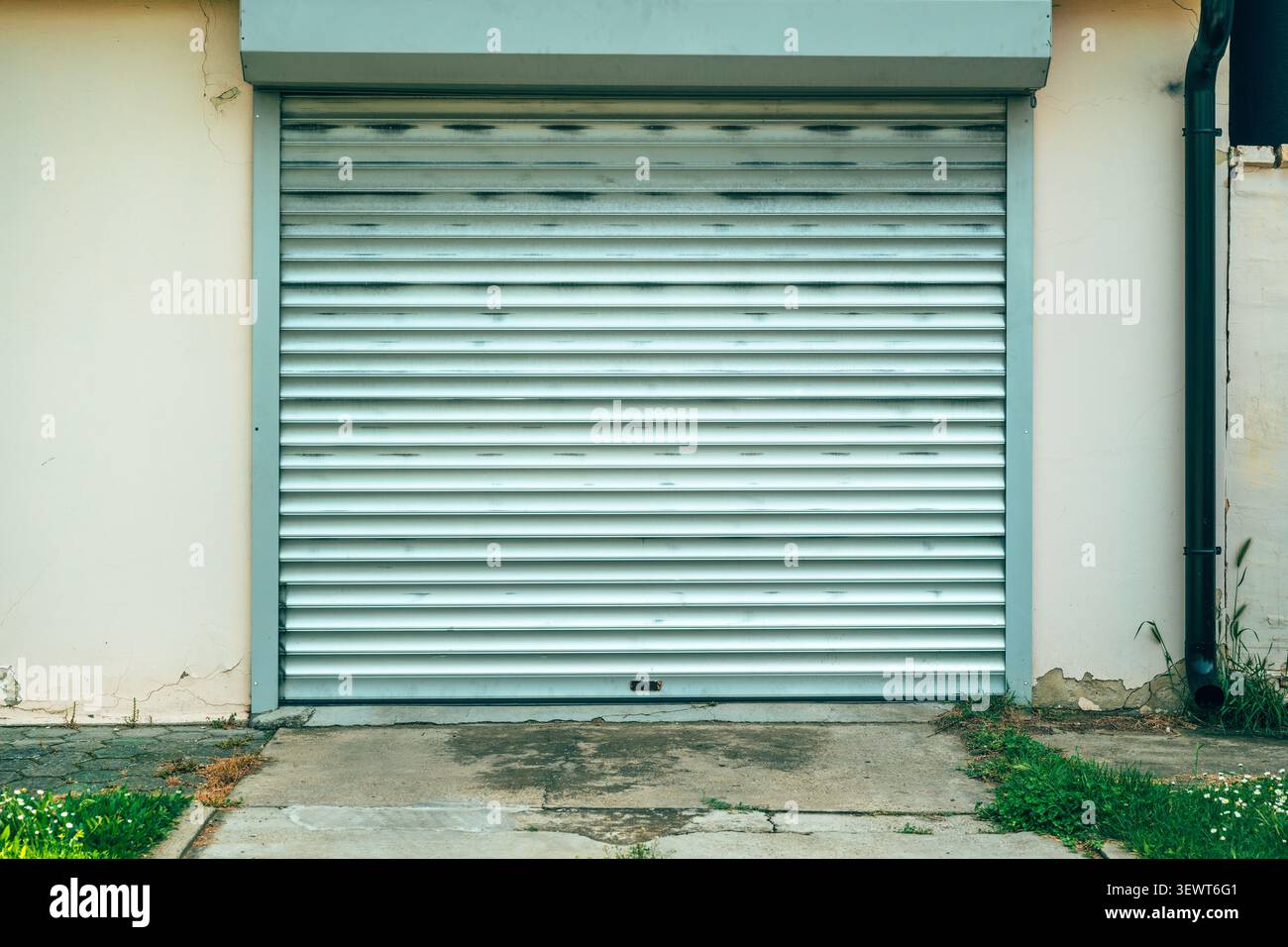La porta del garage in metallo chiusa con pannelli di avvolgimento orizzontali si trova in una parete suburbana intemprata sopra un vialetto di cemento incrinato, incorniciato da uno scarico Foto Stock