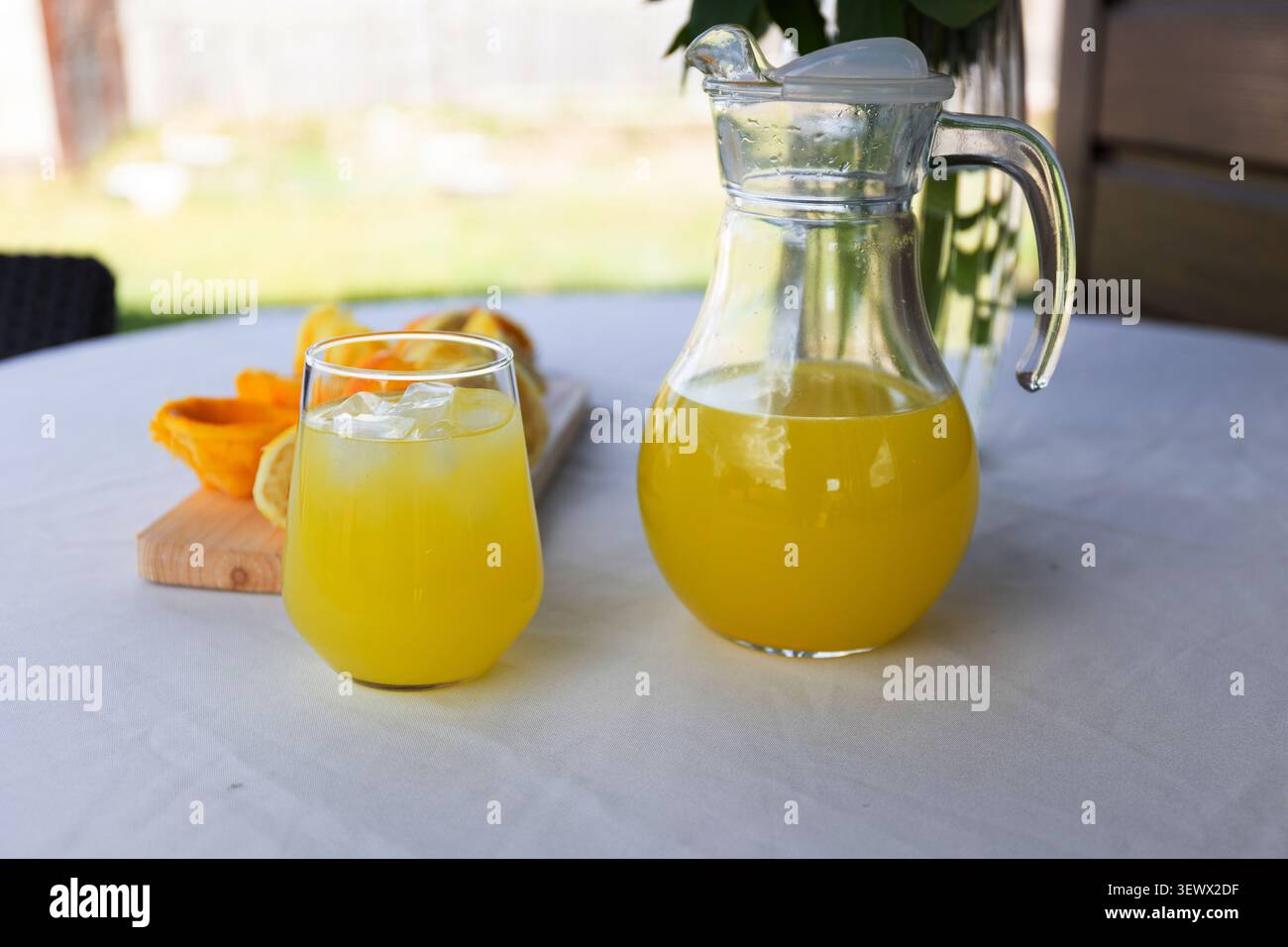 Caraffa in vetro trasparente riempita con succo d'arancia brillante accanto a un bicchiere corto di succo con ghiaccio su tovaglia bianca rotonda, tagliere in legno con arancia a fette Foto Stock