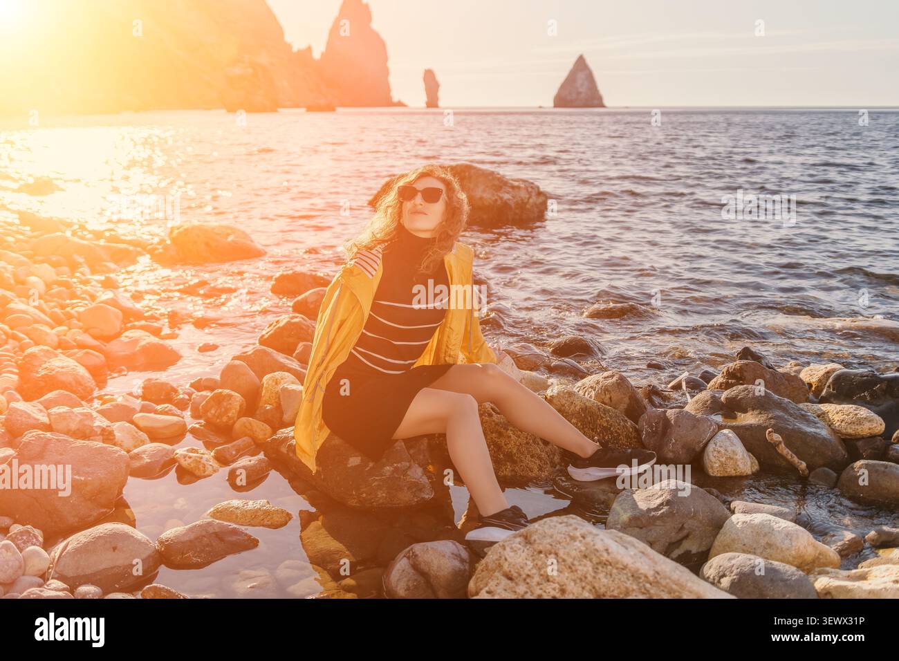 Il tramonto sulla costa delle donne si siede su una spiaggia rocciosa indossando una giacca gialla e occhiali da sole, godendosi una serena serata sulla riva Foto Stock