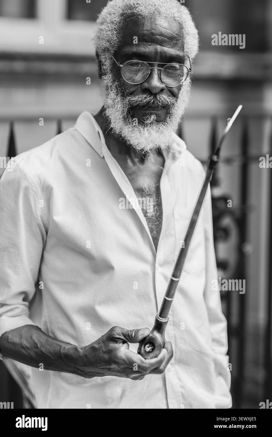 Immagine in bianco e nero di un ritratto di un fumatore di pipa insolito al Windrush Big Caribbean Lunch 2026 a Londra. Foto Stock