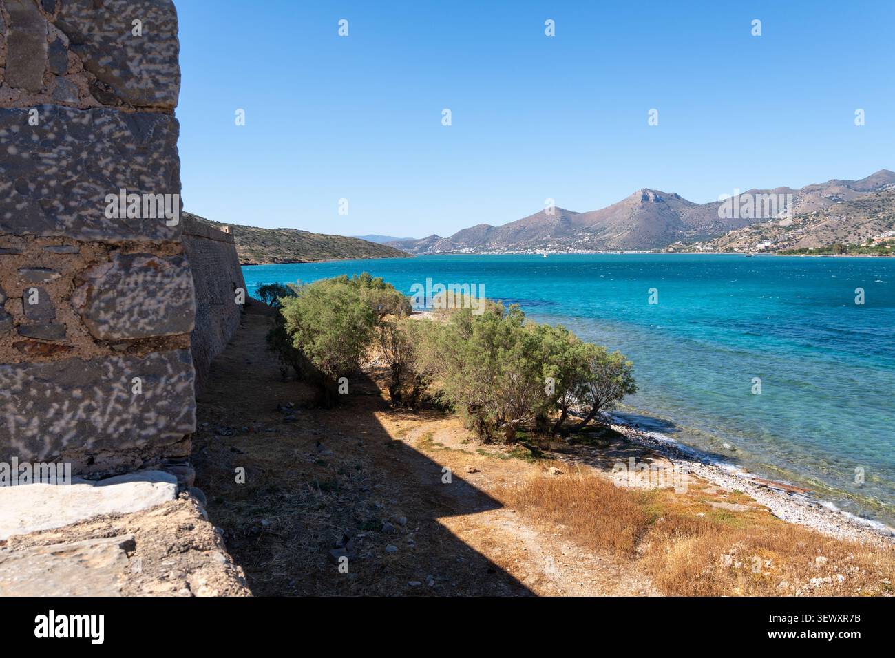 La fortezza di pietra sull'isola di Spinalonga si erge sopra la costa rocciosa con alberi verdi che si affacciano sulle acque turchesi del mare e sulle montagne. Foto Stock