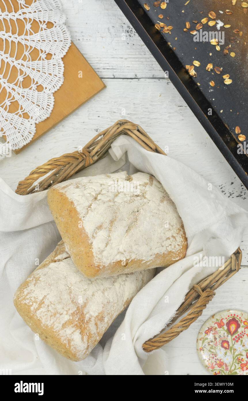 Ingredienti locali, regionali, gusto locale: Panini disposti su un panno di lino nel cestino di vimini. Vassoio marrone, fiocchi d'avena versati e tagliere. Foto Stock