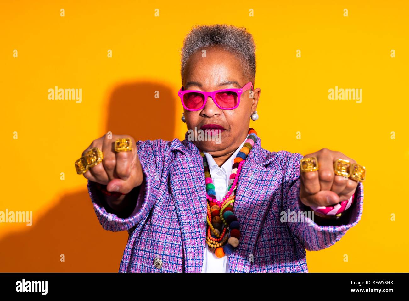 Donna anziana nera che indossa un ritratto di abbigliamento moderno sullo sfondo - donna afroamericana anziana che posa con un atteggiamento sicuro. Indossa occhiali rosa Foto Stock