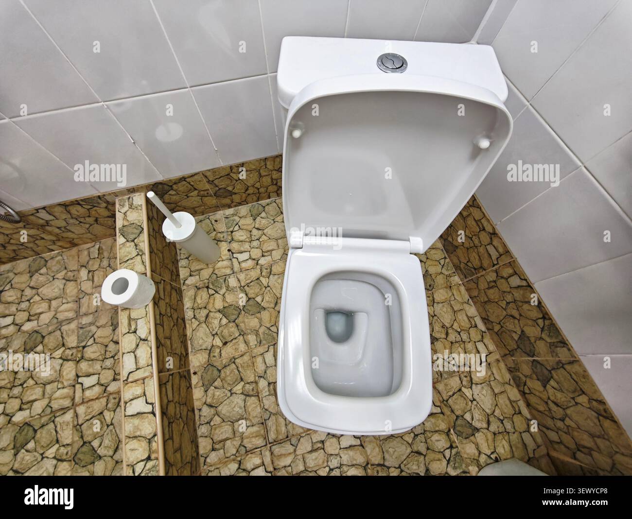 Una toilette bianca si trova in un piccolo bagno con pavimento piastrellato. Presenta un design semplice e pulito. Per comodità, nelle vicinanze sono collocati oggetti di uso quotidiano. Foto Stock