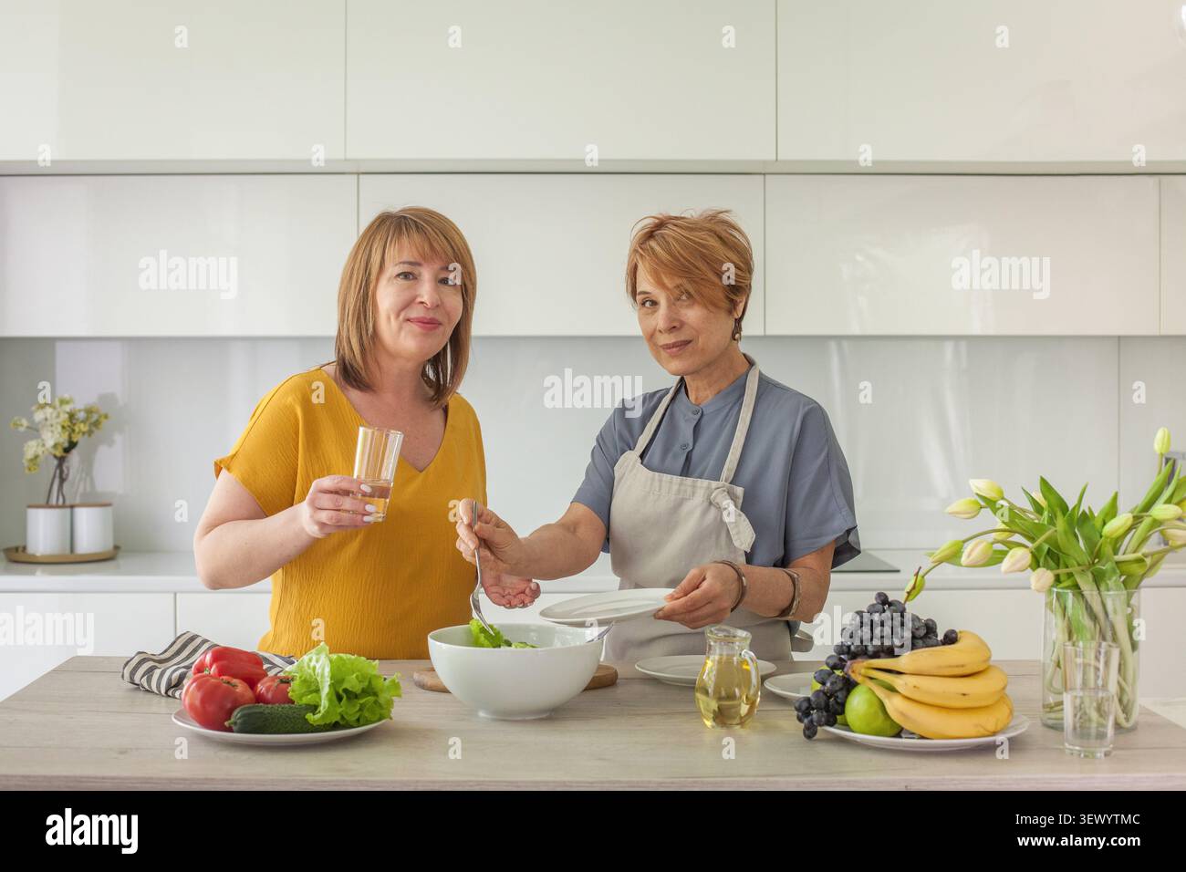 Due donne lavorano insieme in una cucina moderna per preparare un'insalata fresca. Uno contiene un bicchiere di bevanda mentre l'altro mescola gli ingredienti in una ciotola. Loro hanno Foto Stock