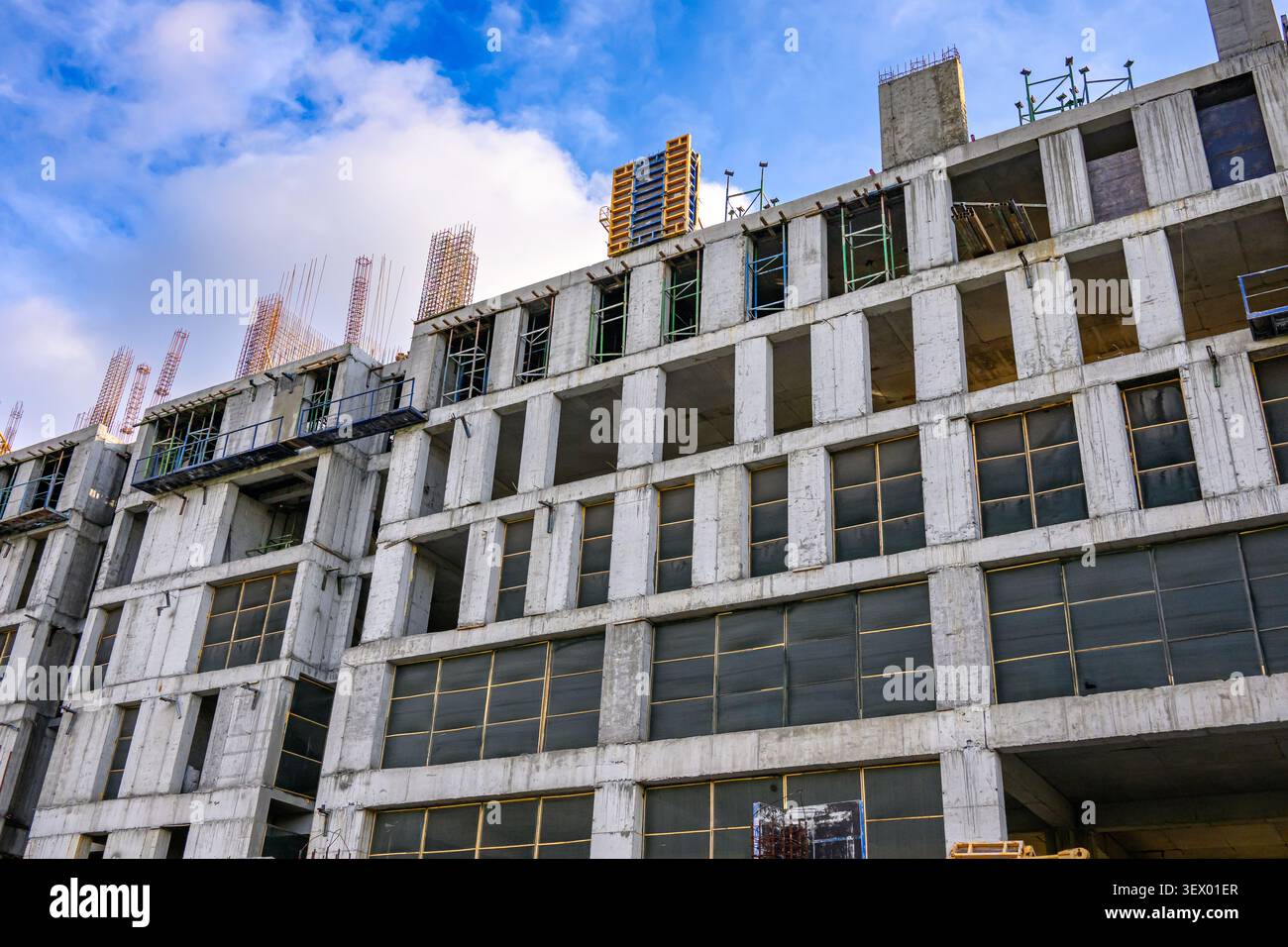 Grande edificio in cemento in costruzione con telai in acciaio visibili e impalcature contro un cielo blu con nuvole sparse Foto Stock