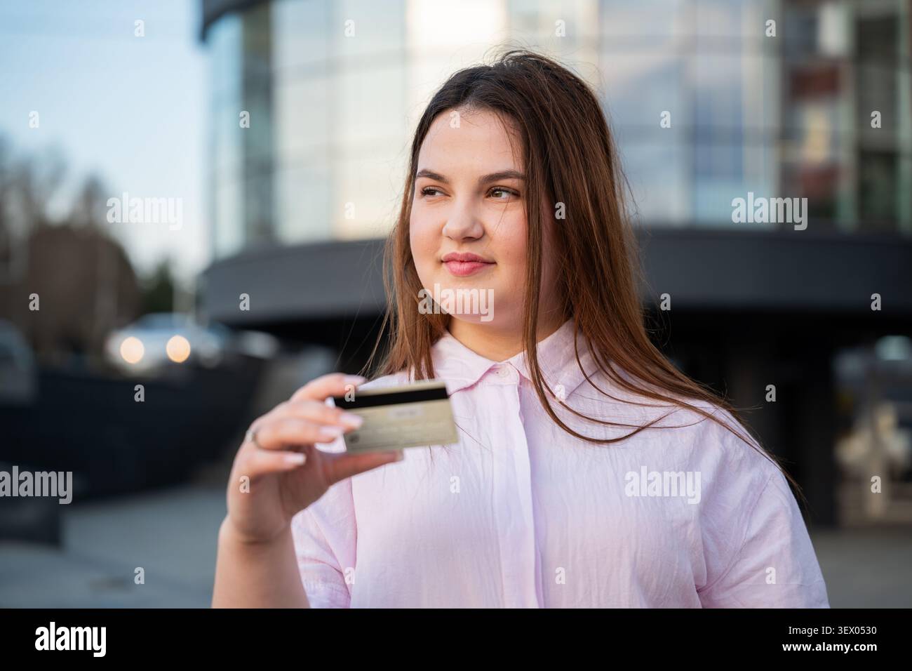 Giovane donna che tiene in mano una carta di credito all'esterno di un moderno edificio per uffici durante la giornata lavorativa. Pagamento online e transazioni digitali sicure per il cashl quotidiano Foto Stock