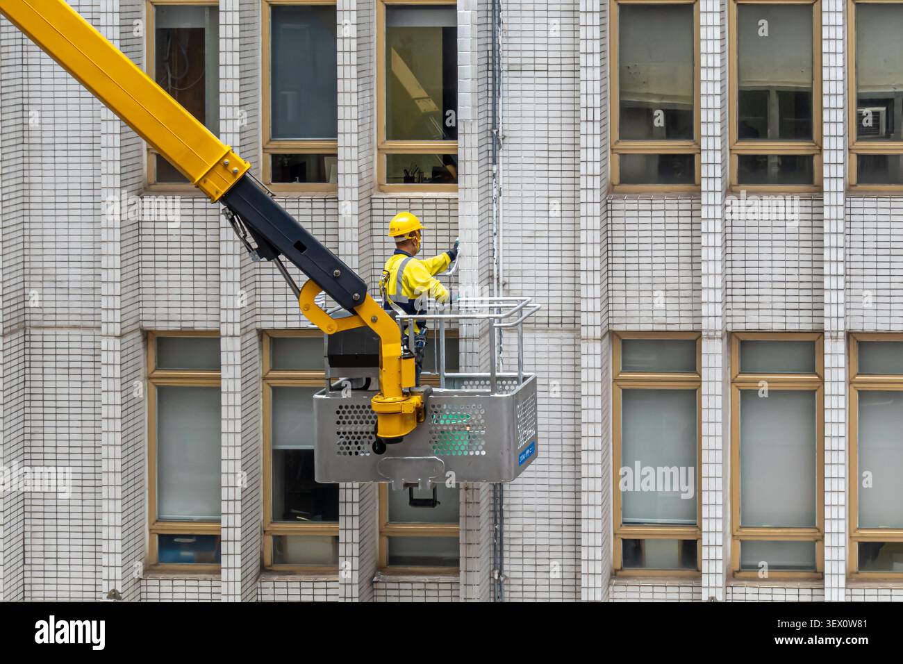 Un lavoratore con un giubbotto ad alta visibilità e un casco giallo in una piattaforma di lavoro mobile elevabile (MEWP), che pulisce la facciata esterna, Tsim Sha Tsui, Hong Kong Foto Stock