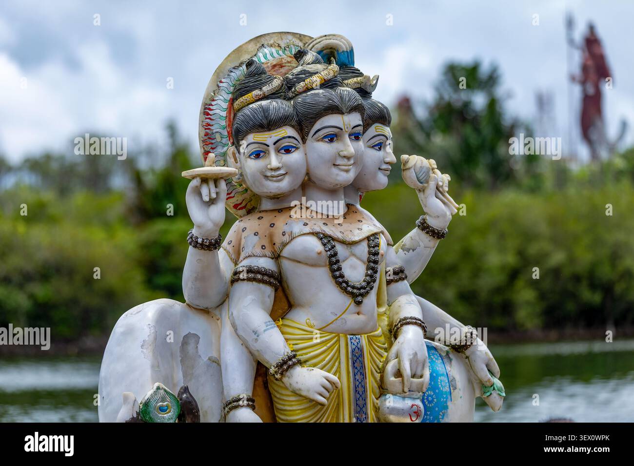 Dettagliata statua di Trimurti con facce e braccia multiple, adagiata su una vegetazione lussureggiante Foto Stock