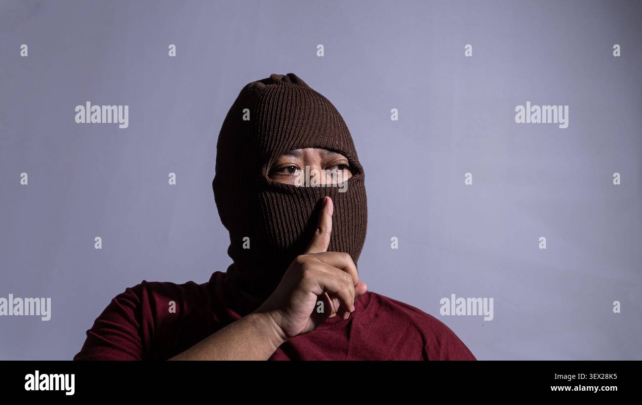 Un maschio adulto che indossa una balaclava a maglia marrone e una camicia marrone tiene un dito sulle labbra coperte con un gesto di silenzio. Guarda di lato Foto Stock