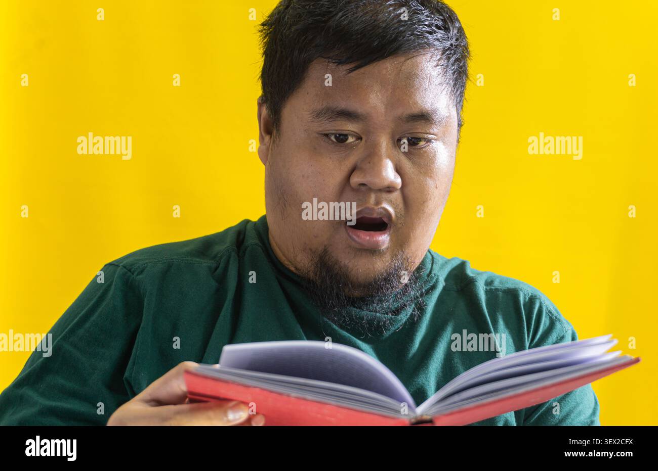Uomo asiatico in camicia verde che legge il libro rosso, reazione sorpresa contro lo sfondo giallo, concetto di scoperta e curiosità nell'apprendimento. Foto Stock