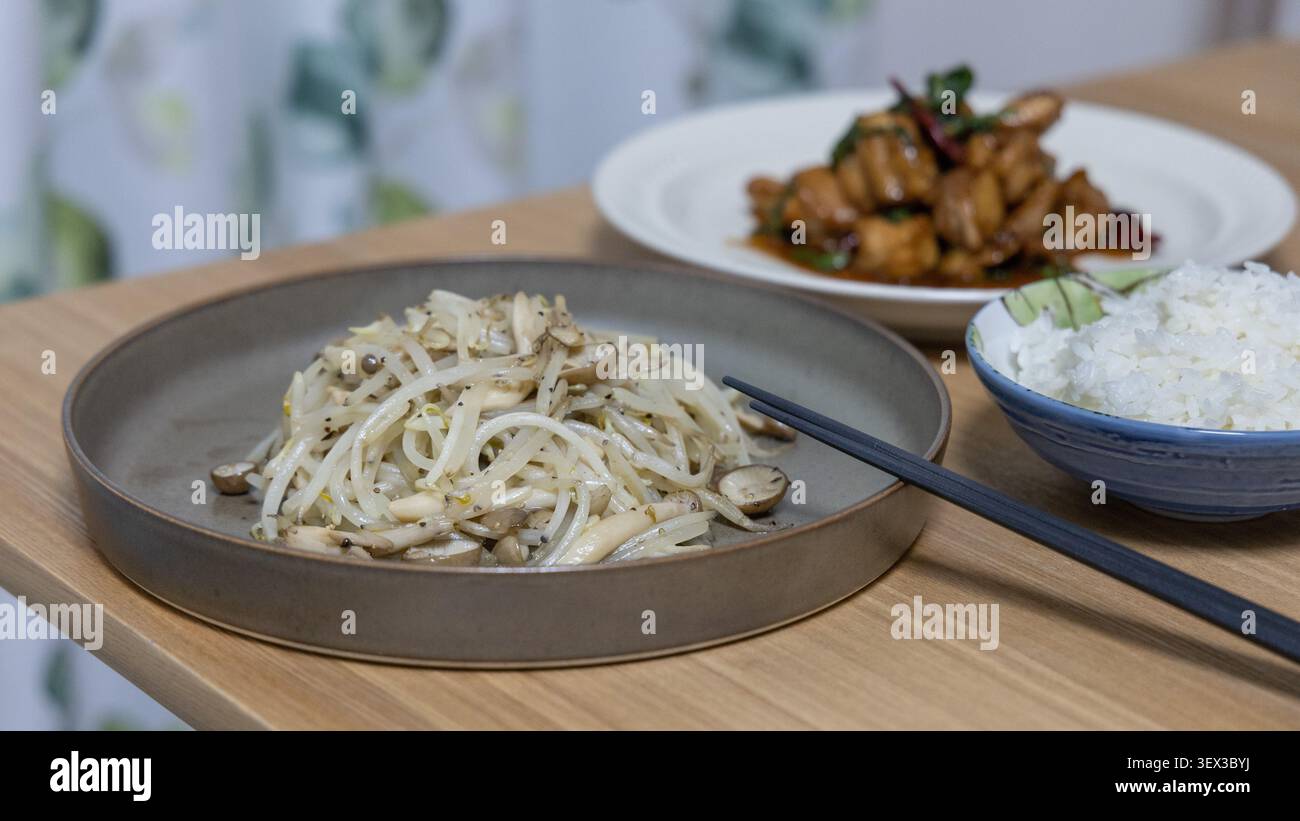 Un pasto cinese fatto in casa, distribuito su un tavolo di legno, presenta germogli di fagioli saltati in padella con funghi in primo piano, tre tazze di pollo e riso al vapore sullo sfondo. Un ambiente familiare caldo e genuino. Foto Stock