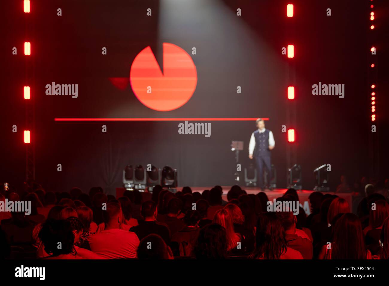 Il relatore sul palco si rivolge al pubblico seduto sotto una spettacolare luce rossa, proiezione del logo visibile sullo sfondo, atmosfera da conferenza, discorso pubblico, evento dal vivo. Foto Stock