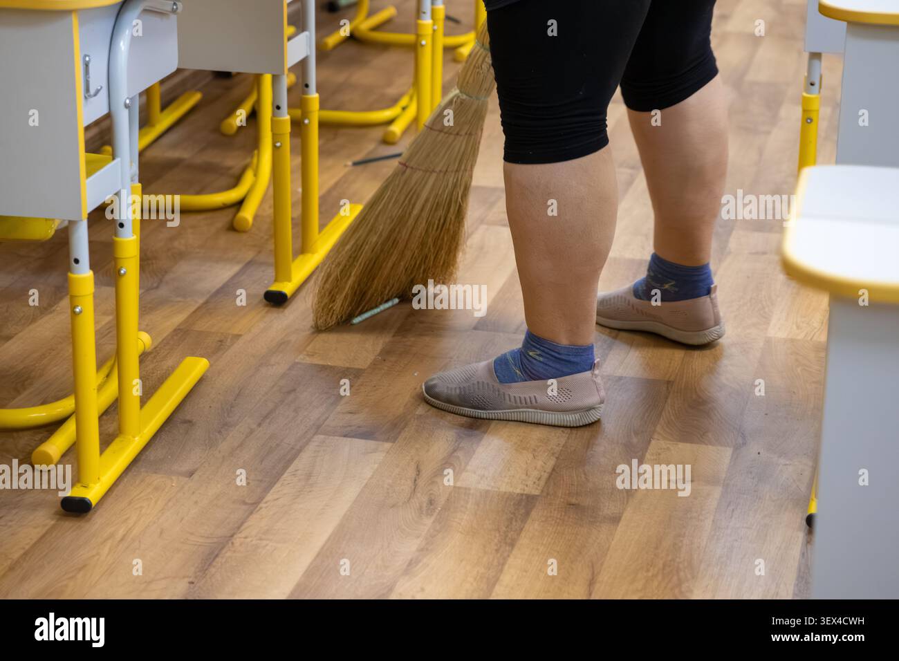 Questo lavoratore dedicato pulisce il piano dell'aula con una scopa vicino alle scrivanie degli studenti per illustrare la manutenzione dell'igiene professionale e i dirigenti della struttura Foto Stock
