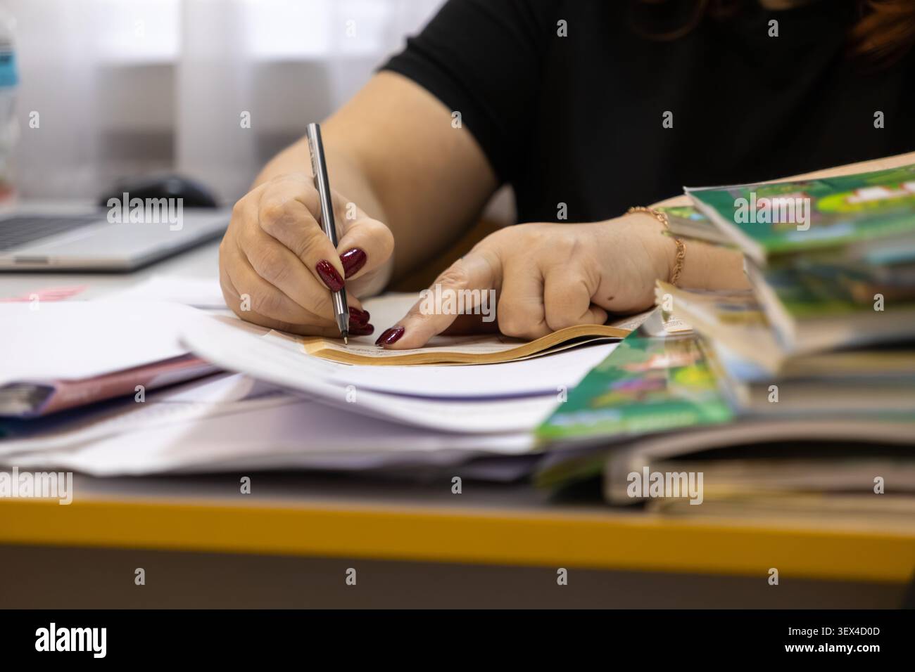 Un individuo concentrato prende appunti in un documento a una scrivania occupata, dove pile organizzate di documenti cartacei e una configurazione al computer illustrano un approccio dedicato Foto Stock