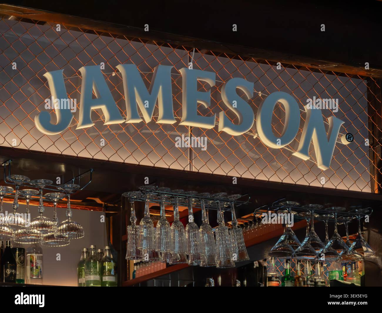 Kiev, Ucraina - 21 marzo 2026: Marchio Jameson luminoso dietro uno schermo in rete metallica sopra un bar taverna dove file di bicchieri da cocktail puliti ha Foto Stock