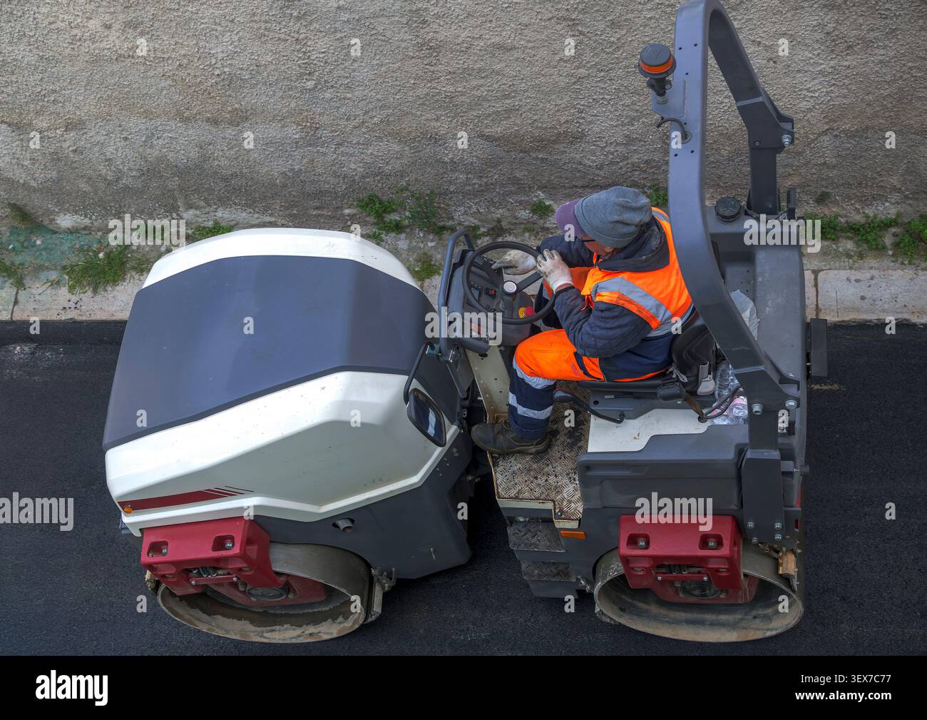 Vista ad angolo alto di un rullo compatitore vibrante a doppio tamburo che compatta attivamente asfalto nero fresco durante la costruzione di una strada, azionato da un industriale Foto Stock