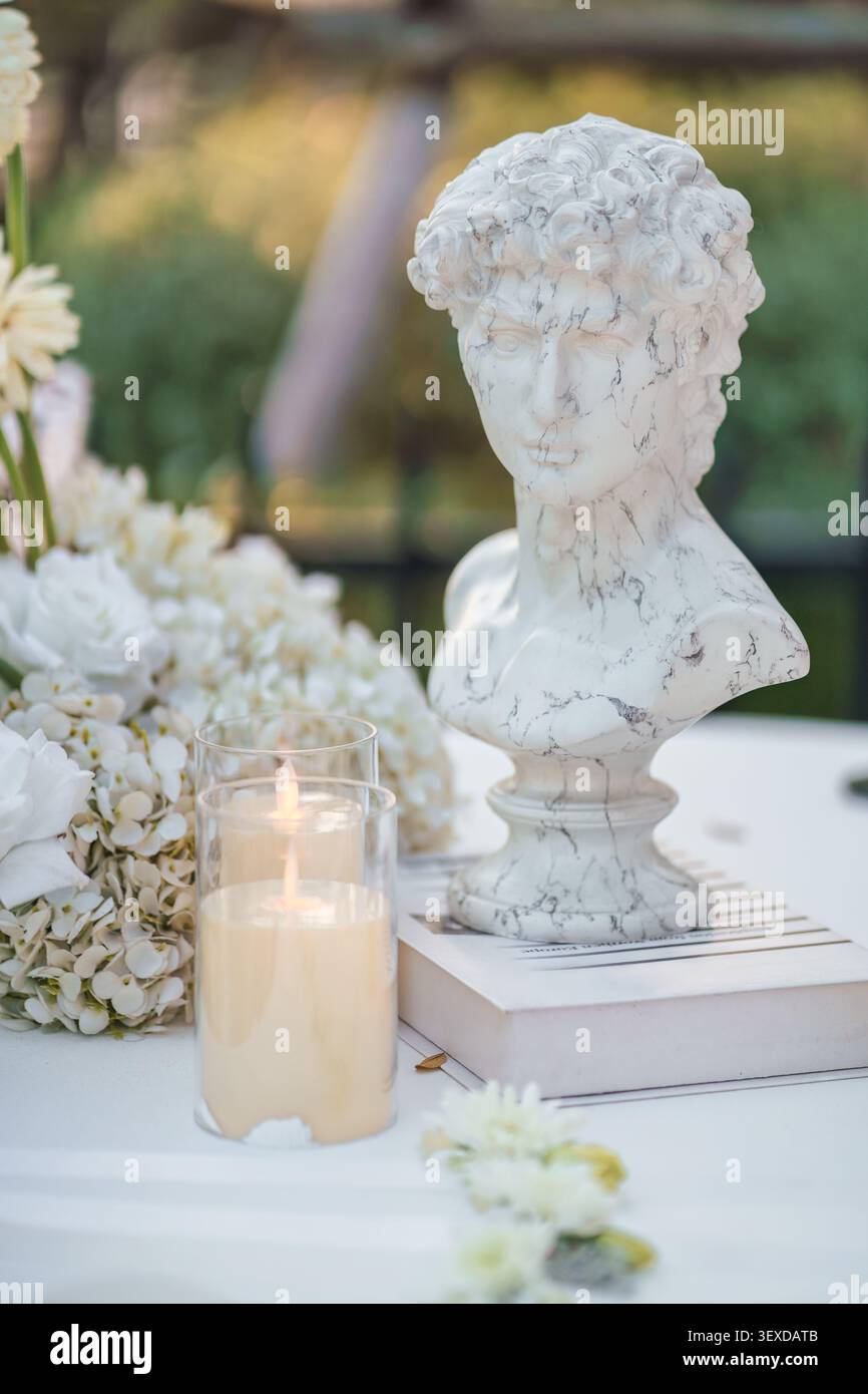 La donna classica affronta una scultura in marmo sul libro con candela e fiori su un tavolo decorativo per temi di arte, design, matrimonio e celebrazione Foto Stock