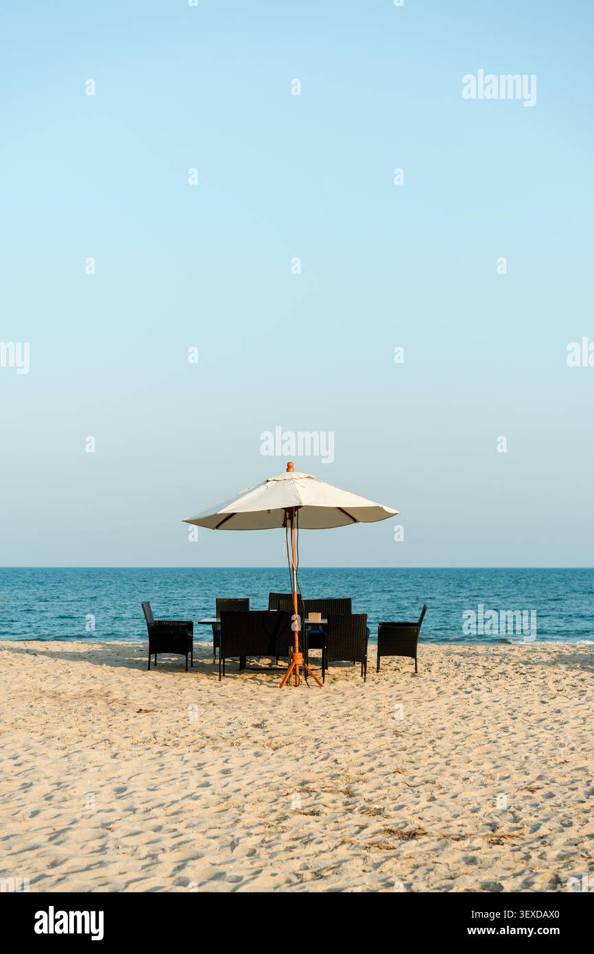 Elegante tavolo da pranzo con sedie in vimini e ombrellone bianco sotto il cielo limpido sulla spiaggia di sabbia durante il pomeriggio Foto Stock