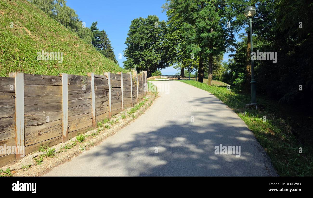 Un sentiero asfaltato si snoda verso l'alto, accanto a una barriera in legno che regge una collina erbosa, fiancheggiata da lussureggianti alberi verdi e da un classico semaforo Foto Stock