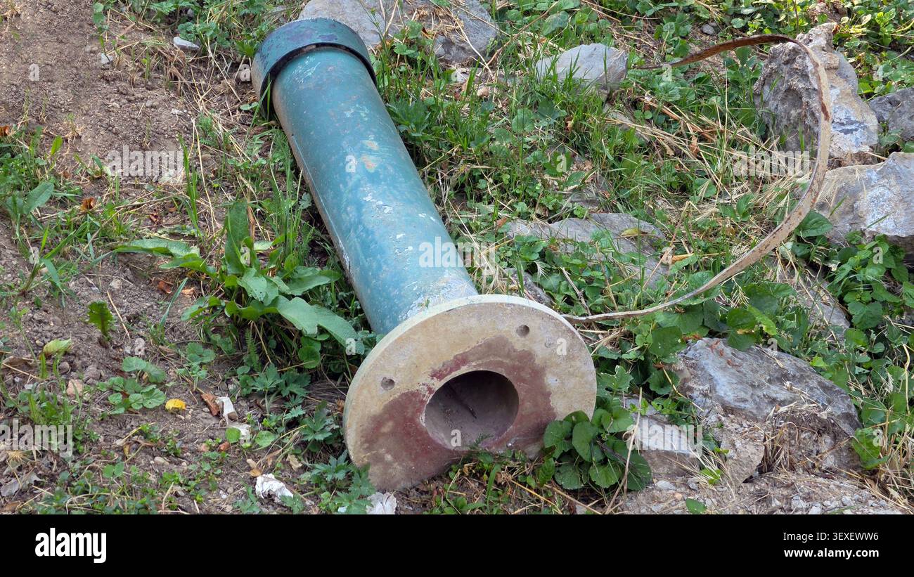 Una barriera cilindrica scollegata in acciaio poggia su un terreno sterrato e erboso circondato da pietre sparse e erbacce selvatiche Foto Stock