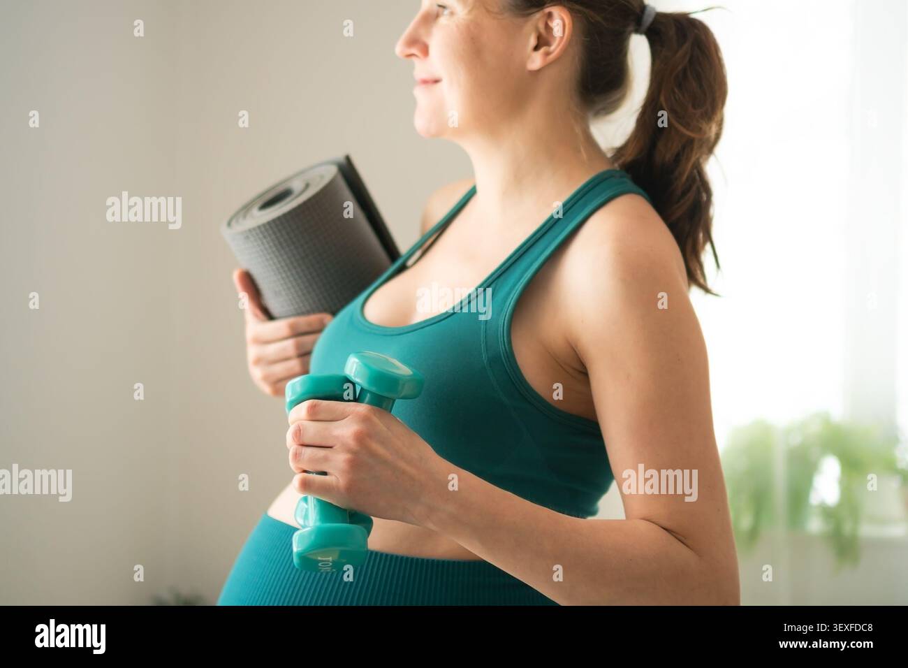 Donna incinta in abbigliamento sportivo turchese che si esercita in sicurezza a casa. Aspettatevi che la mamma tenga il manubrio e il tappetino per lo yoga per un allenamento attento e delicato. La mamma-a-essere rimane protetta dando priorità a una gravidanza sana Foto Stock