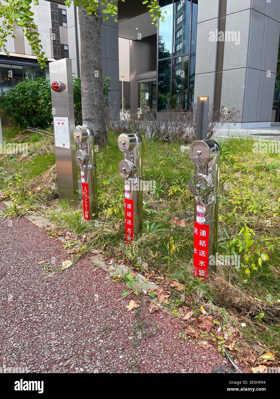 Primo piano del moderno sistema di collegamento degli idranti antincendio in acciaio inossidabile con etichette rosse nella strada cittadina giapponese, circondato da piante verdi. Fukuoka, Giappone Foto Stock