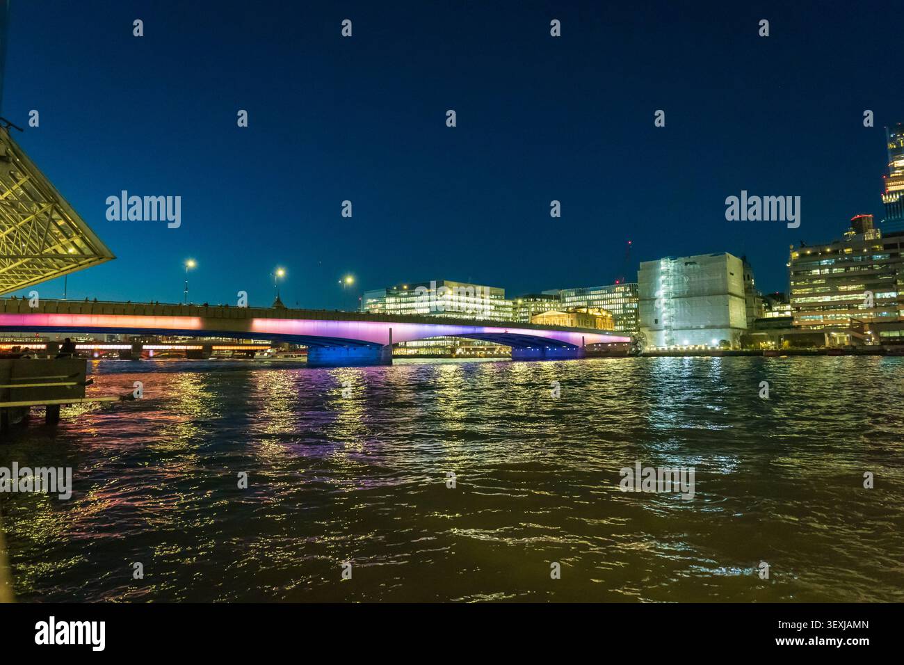 Il lungofiume della città notturna con un ponte illuminato e moderni edifici per uffici che proiettano riflessi caldi sull'acqua ondulata, evocando la calma urbana, eveni Foto Stock