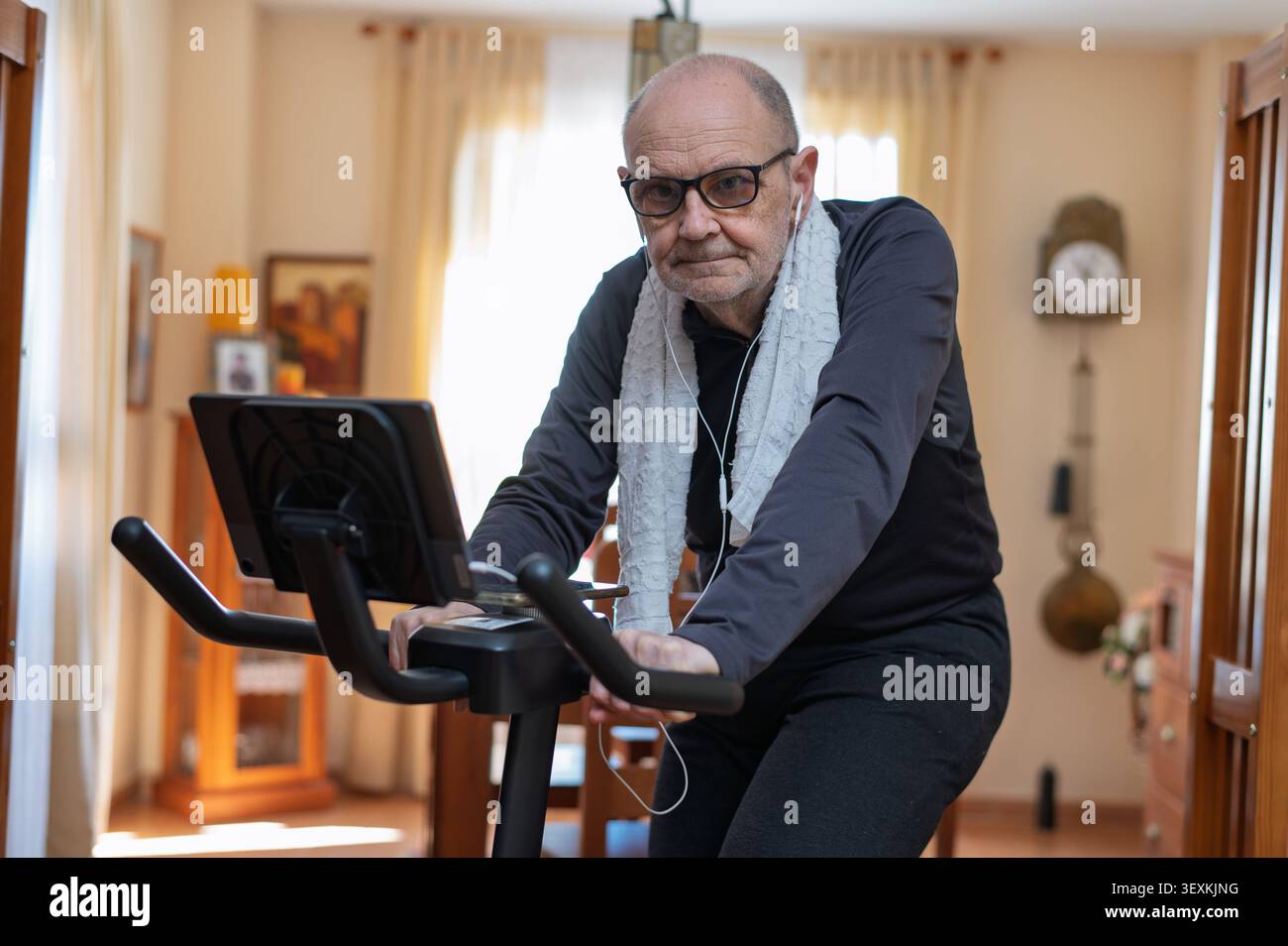 Un anziano pedala una cyclette nel suo salotto, indossando cuffie e un asciugamano, concentrandosi sulla forma fisica a basso impatto e sull'invecchiamento sano con una Foto Stock