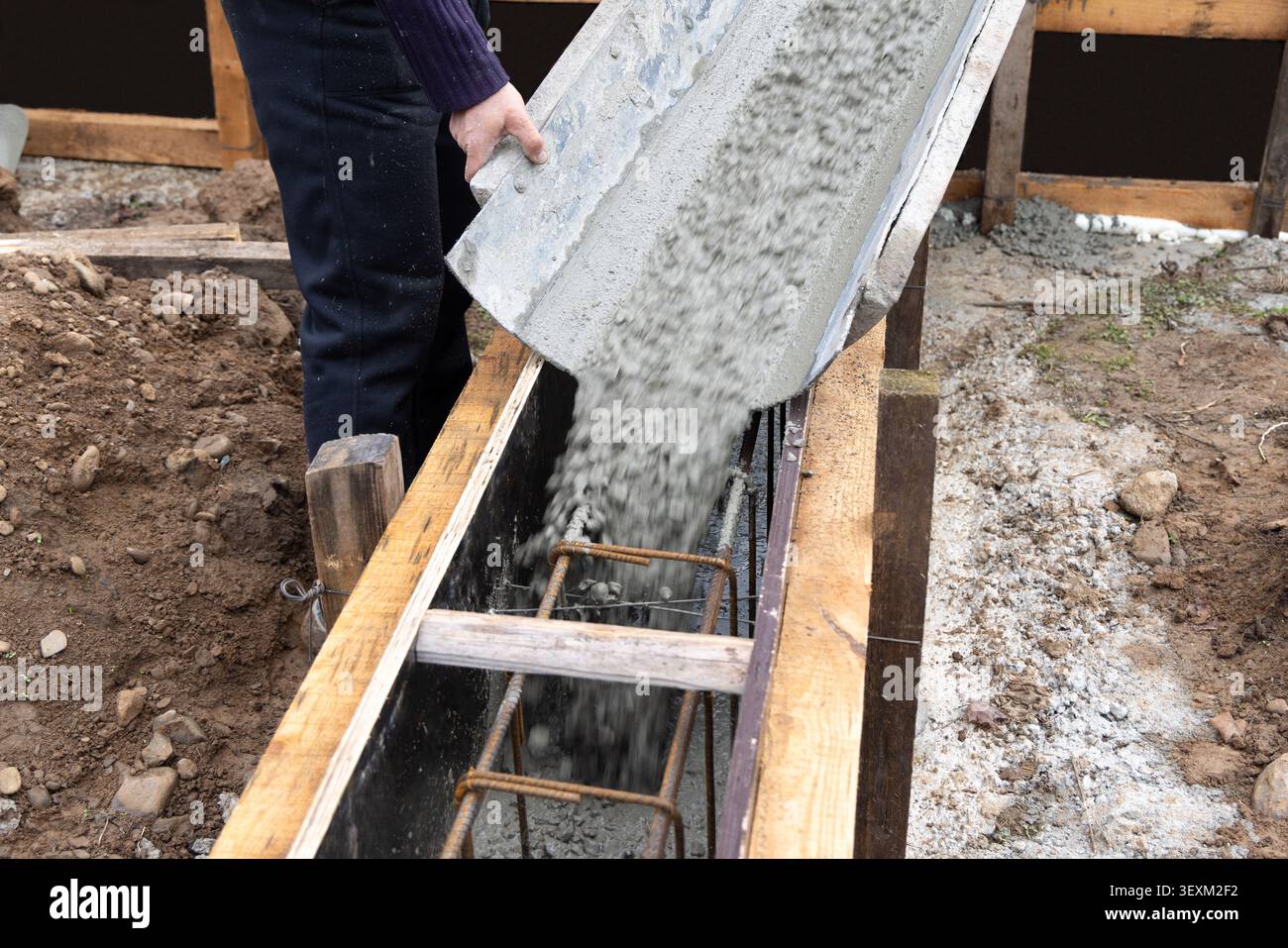 Il calcestruzzo viene versato in casseforme di legno con rinforzo in acciaio nel cantiere. Operatore che guida il flusso della miscela di cemento fresco durante la fondazione Foto Stock