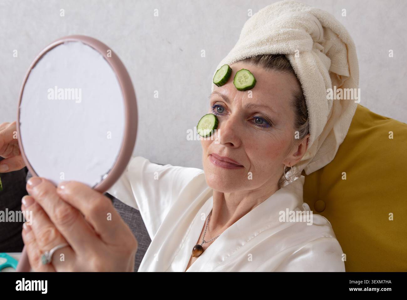 La donna anziana si siede comodamente sul divano avvolto in un asciugamano, applicando la cura della pelle con l'aiuto di fette di cetriolo, appoggiandosi sulla fronte, l Foto Stock